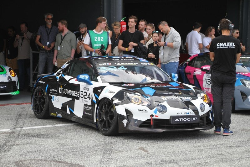 Johan Akkerman's Alpine A110 Cup race car number 324 in the paddock with team members, friends and supporters in the background.